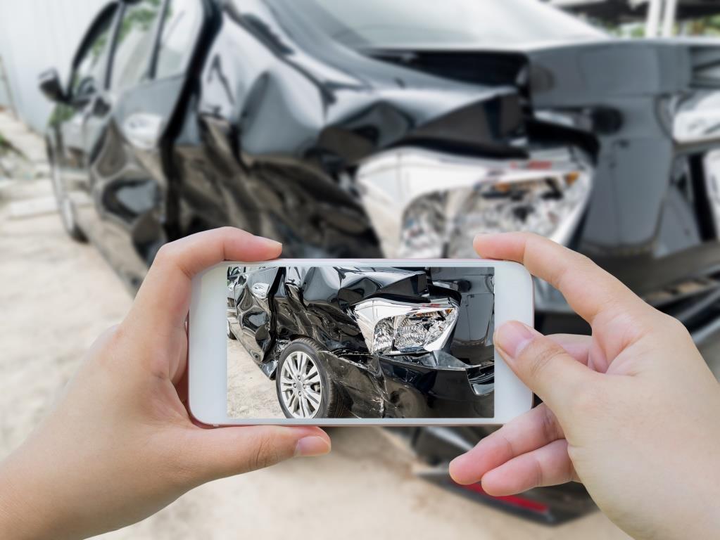 Person holding a smartphone and taking a photo of a damaged car. The car has significant damage to its rear, with visible dents and crumpled metal. The focus of the smartphone camera is on the damaged area, capturing the details of the accident. This scenario likely depicts the documentation of car damage for insurance purposes.