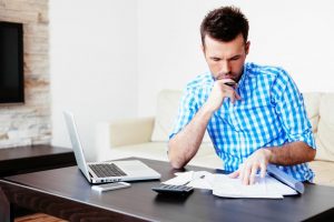 Person sitting in front of an open laptop, examining some papers in front of him. Also has calculator and phone on desk