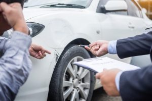 An insurance adjuster and a car owner examining the damage on a white vehicle. The adjuster is pointing at the damage with a pen and holding a clipboard, while the car owner is gesturing towards the damaged area near the front wheel.